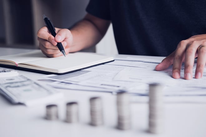 A person reviewing documents and taking notes with stacks of money in the foreground