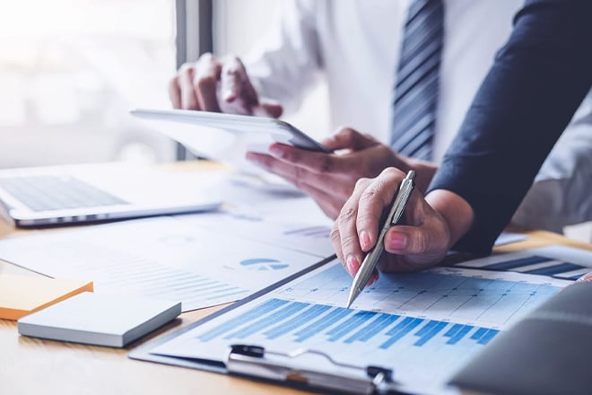 Close up of two coworkers in business attire reviewing data on documents and screens