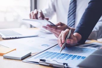 Close up of two coworkers in business attire reviewing data on documents and screens