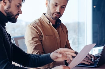 Two men are seated at a desk looking at printed documents. One man is pointing at the papers while the other is focused on the discussion.