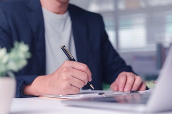 Close-up of a businessman writing on a clipboard while looking at his laptop.