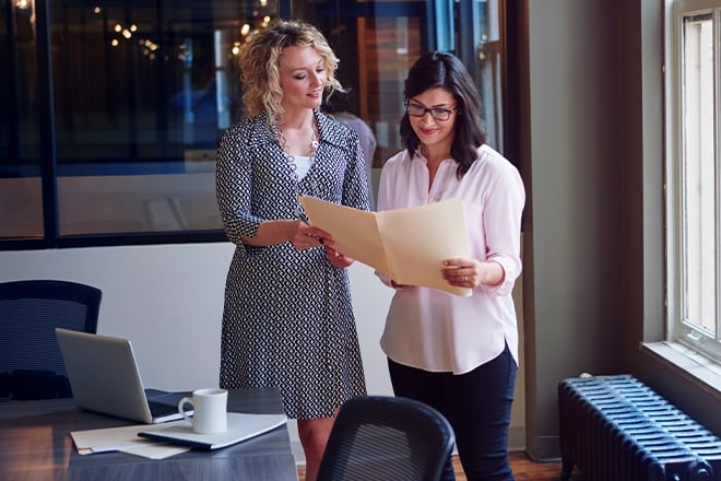 Two women holding a folder open to look at the documents inside while standing in a conference room.