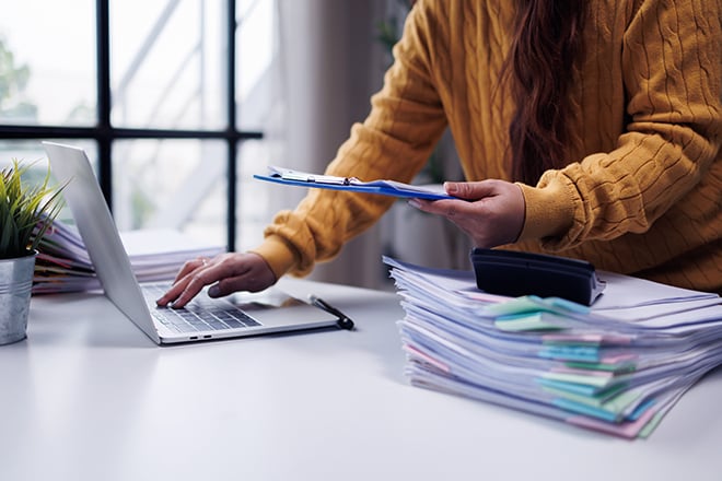A woman using a laptop next to stacks of colorful paperwork