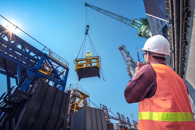 An image of a construction worker on the ground communicating with his co-worker in a crane by walkie talkie