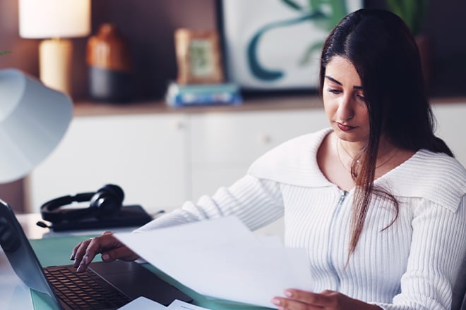 A woman sitting at her desk holding paperwork in one hand while typing on her laptop with the other hand.