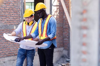 Two construction workers in safety gear reviewing blueprints on site