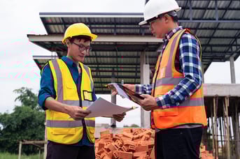 Two construction workers in safety vests and hard hats review documents and use a tablet at a building site.