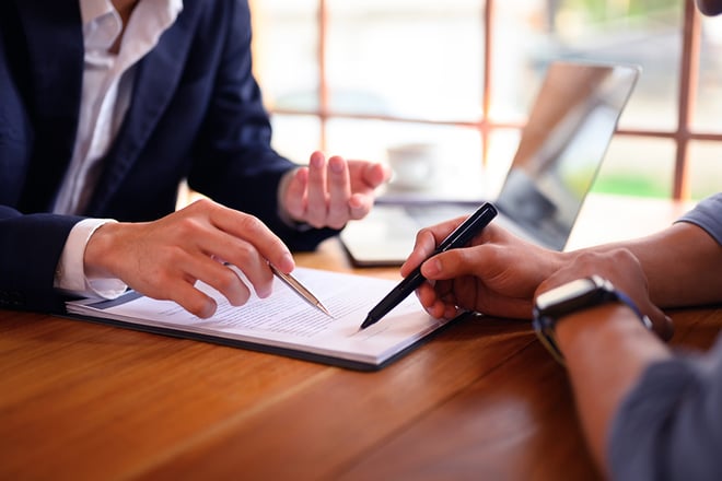Two business professionals reviewing and signing documents at a wooden table with laptop in background.