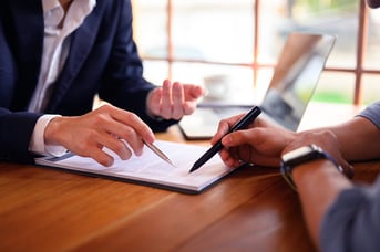 Two business professionals reviewing and signing documents at a wooden table with laptop in background.
