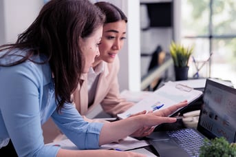 Two women collaborate at a desk, reviewing charts on a laptop and tablet in a bright modern office setting.