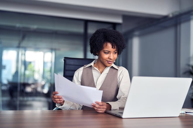 A professional woman with curly hair reviews documents at her desk with a laptop in a modern office setting.