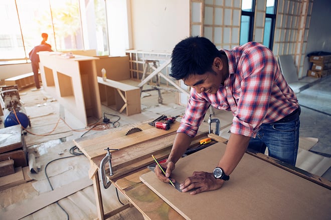 A craftsman in a plaid shirt measures and marks wood with a pencil in a bright workshop filled with tools and materials.