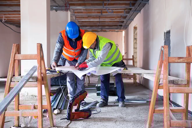 Two construction workers in hard hats and high-visibility vests reviewing blueprints together at an active construction site, surrounded by wooden scaffolding and building materials with exposed ceiling beams visible above