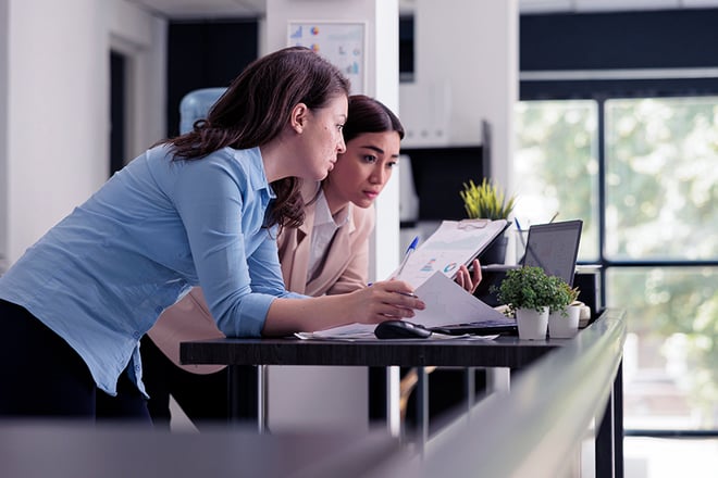 Two women collaborating at a desk, reviewing documents and working on a laptop in a modern office.