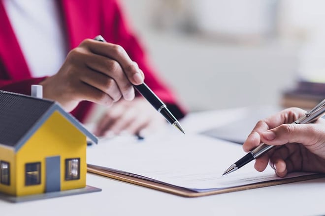 Close up of insurance documents on a desk with two people pointing towards it with pens. There is a miniature home model in the foreground.