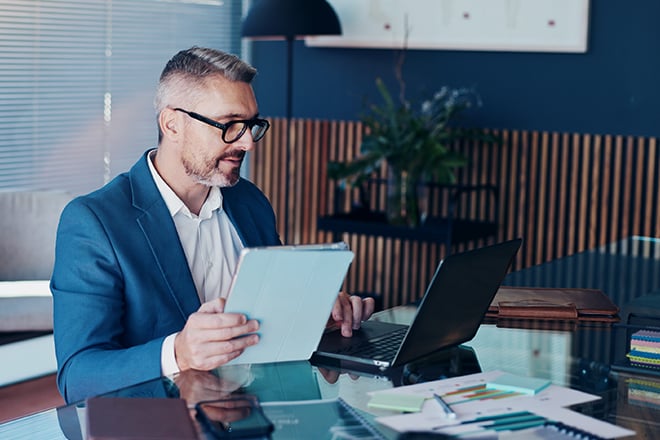 Risk manager in suit reviewing documents on tablet and laptop at office desk