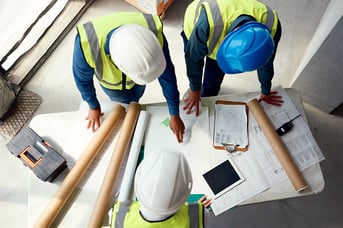 Three construction workers in safety vests and hard hats review blueprints and plans on table at job site.