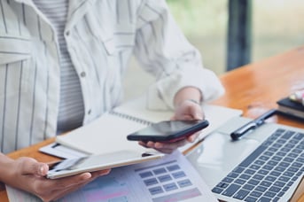 Person in striped shirt comparing products on smartphone and tablet at desk with laptop and calculator.