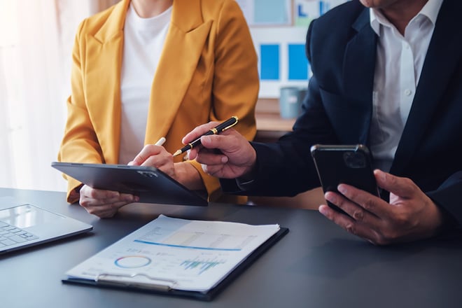 Two business professionals in suits review documents and use devices at a conference table with charts visible.