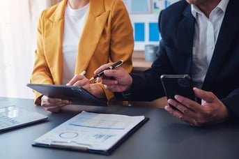 Two business professionals in suits review documents and use devices at a conference table with charts visible.