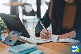woman writing in notebook and looking at tablet at desk