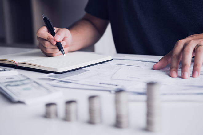 A person reviewing documents and taking notes with stacks of money in the foreground