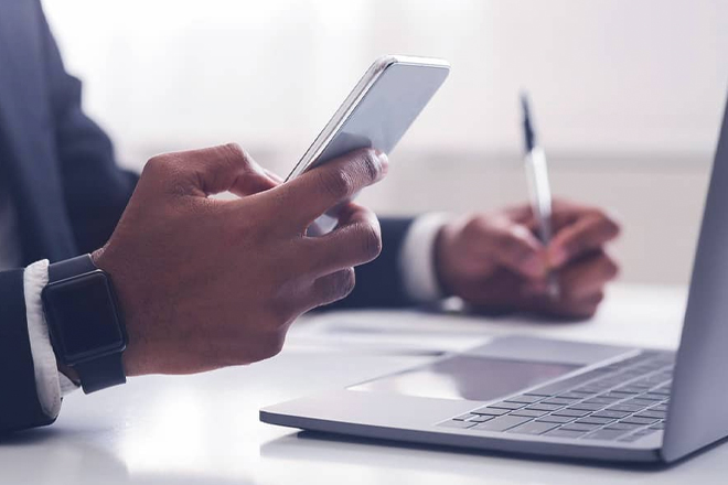 Person using smartphone while working on laptop at desk