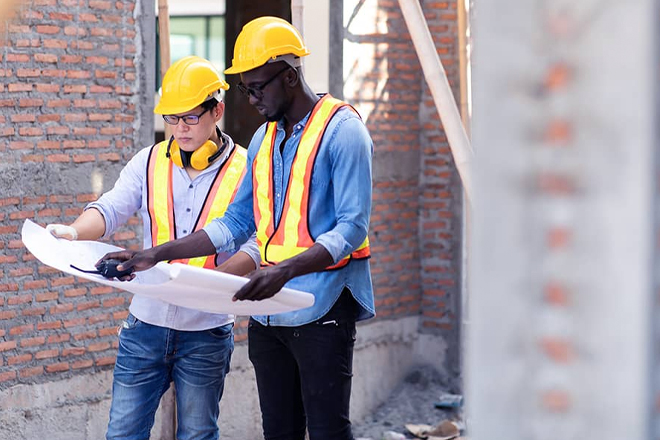 Two construction workers in safety gear reviewing blueprints on site