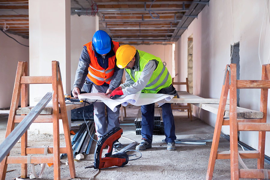 Two construction workers in hard hats and high-visibility vests reviewing blueprints together at an active construction site, surrounded by wooden scaffolding and building materials with exposed ceiling beams visible above