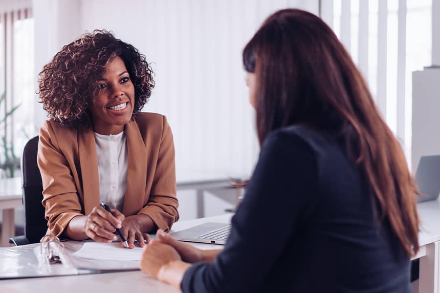 Two women looking at paperwork together at a desk in an office setting. One of them is facing the camera the other has their back to the camera.